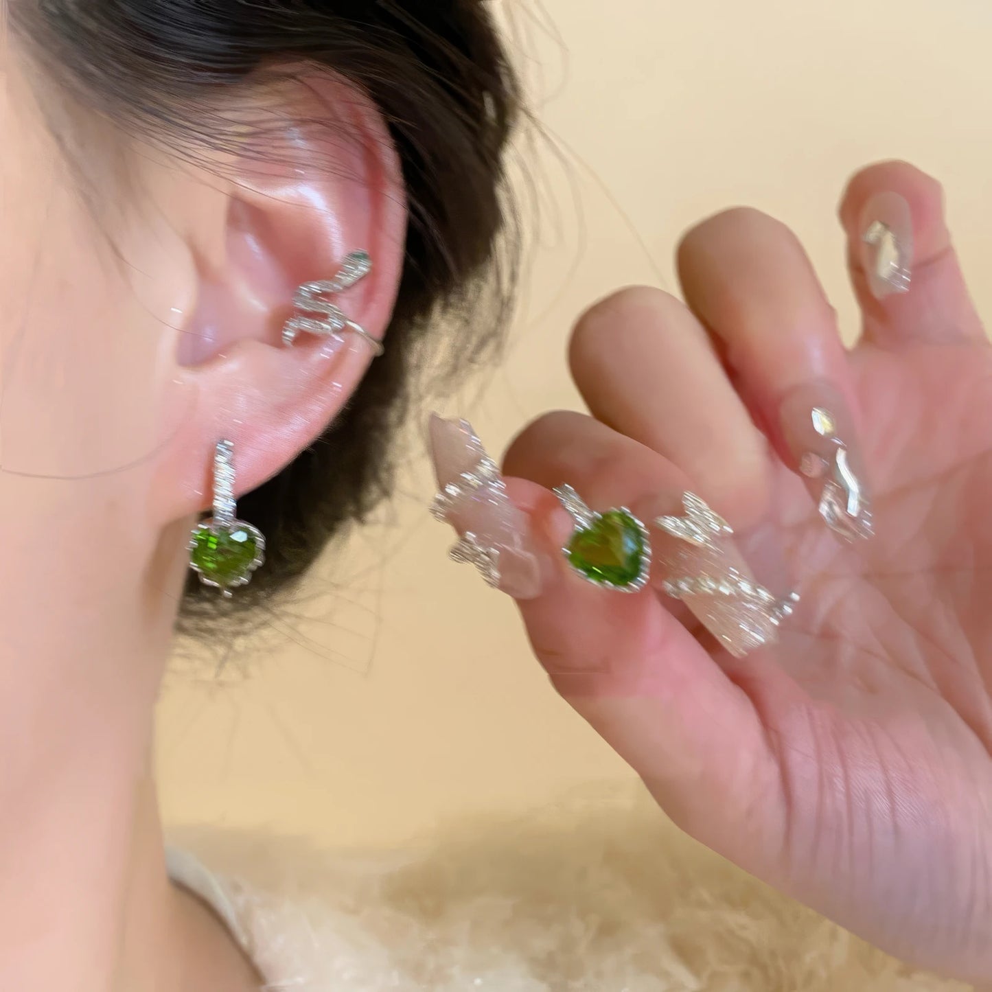 Green zircon snake - shaped stackable ear cuff worn on ear, with a hand holding a matching accessory.