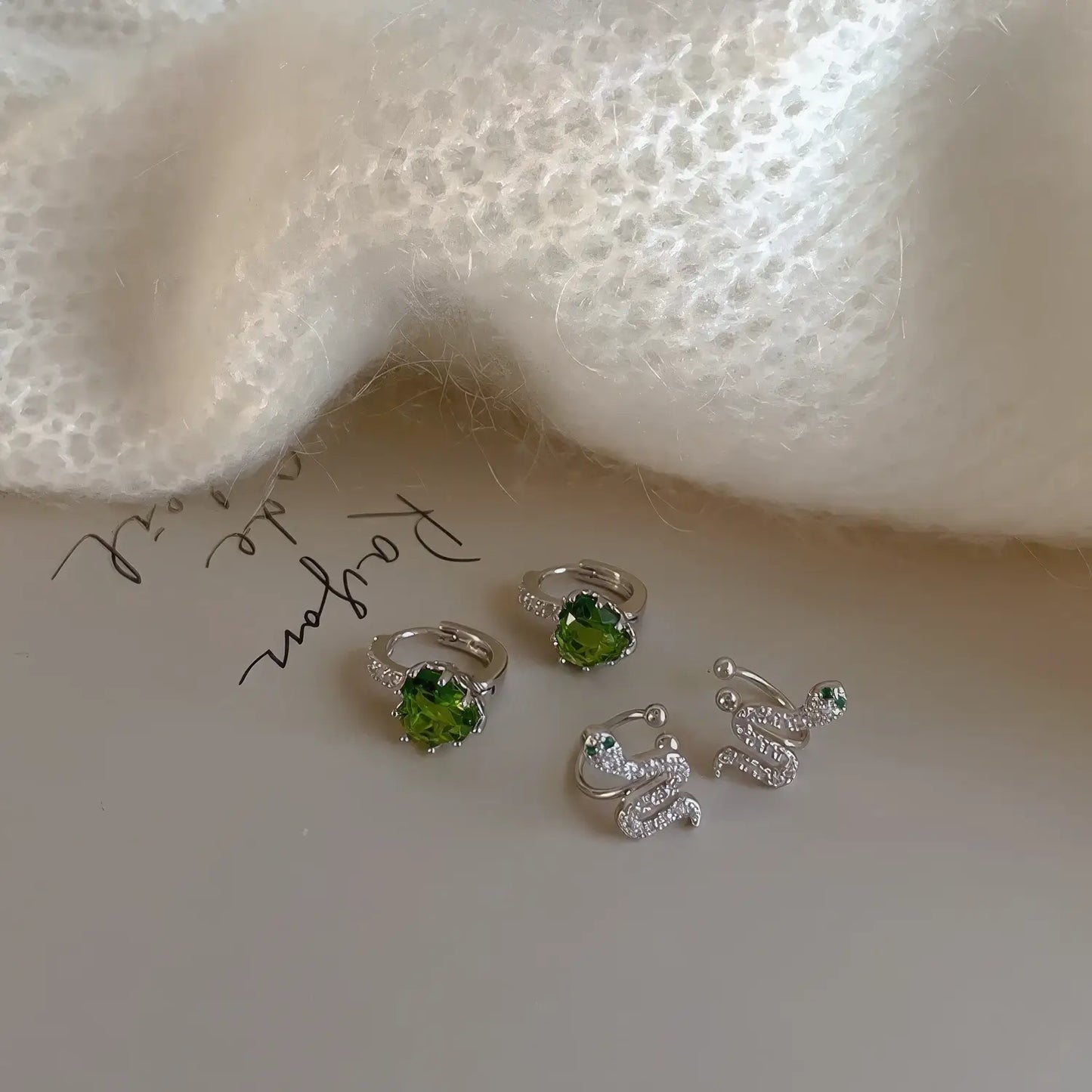 Green zircon snake - shaped stackable ear cuff along with other jewelry pieces, placed on a surface with a knitted fabric.