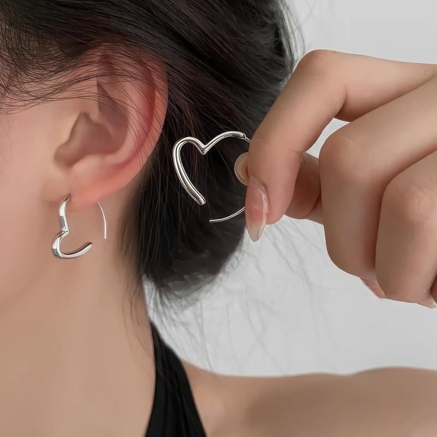 Silver heart - shaped earring worn, with hand holding the other earring, highlighting the delicate, open - heart structure