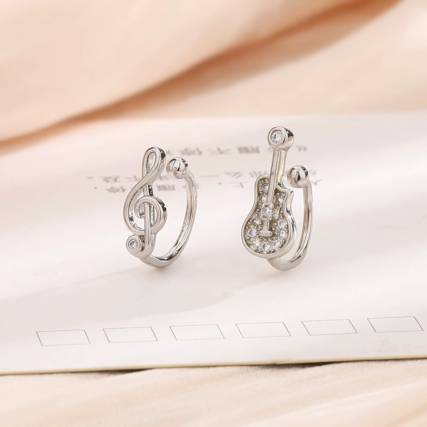 Silver treble clef and guitar - shaped ear cuffs displayed on a light - colored paper, emphasizing the zircon - encrusted musical designs