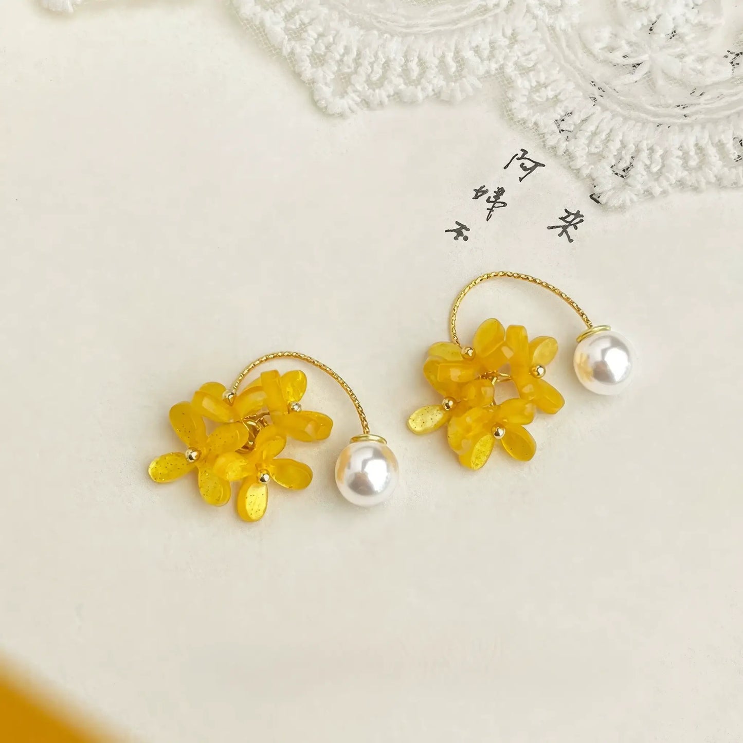 Pair of yellow acrylic floral earrings with pearl accents, displayed on a lace - detailed surface, showcasing the vibrant flower and elegant pearl design