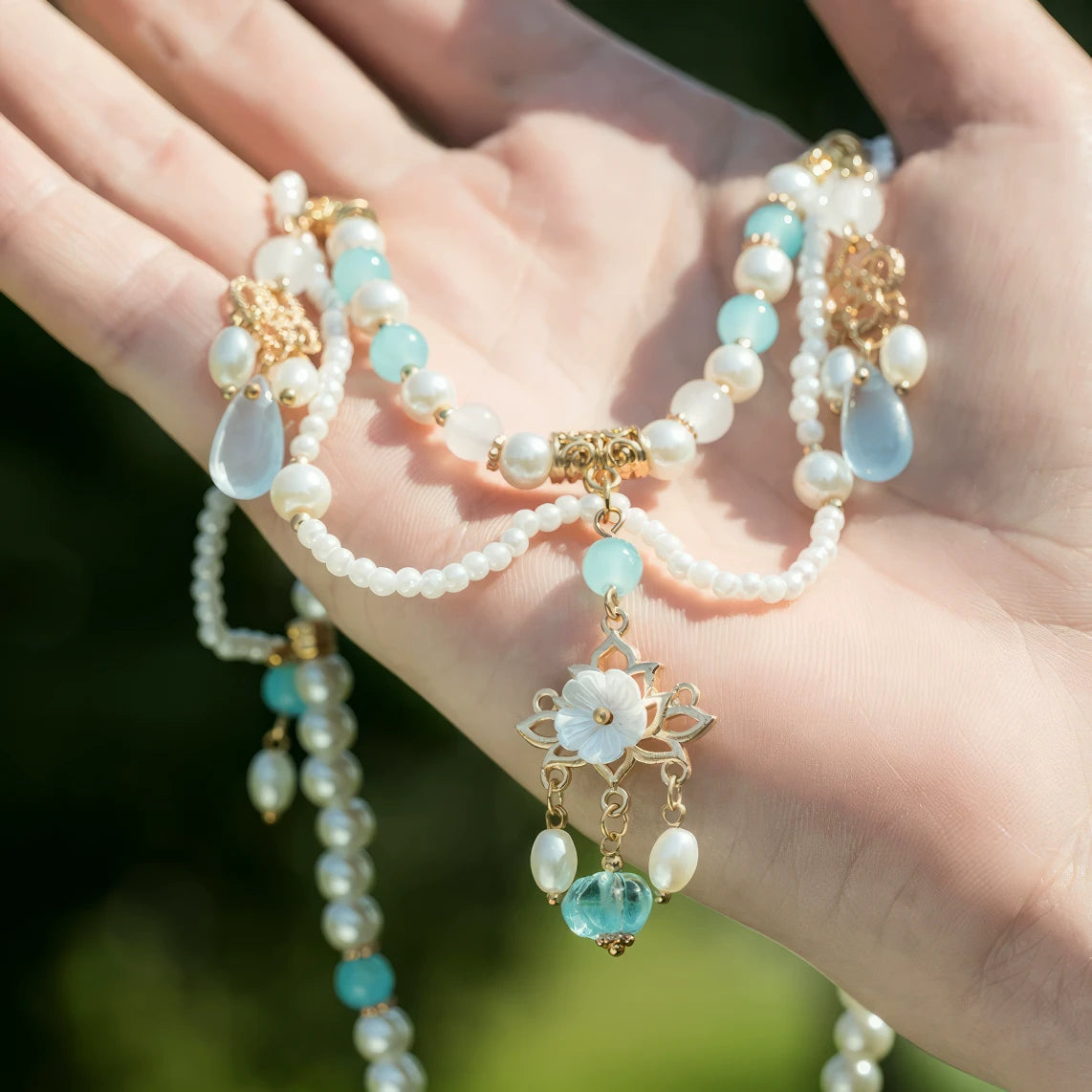 Hand holding blue double - layer necklace with pearls and gold details, outdoor background