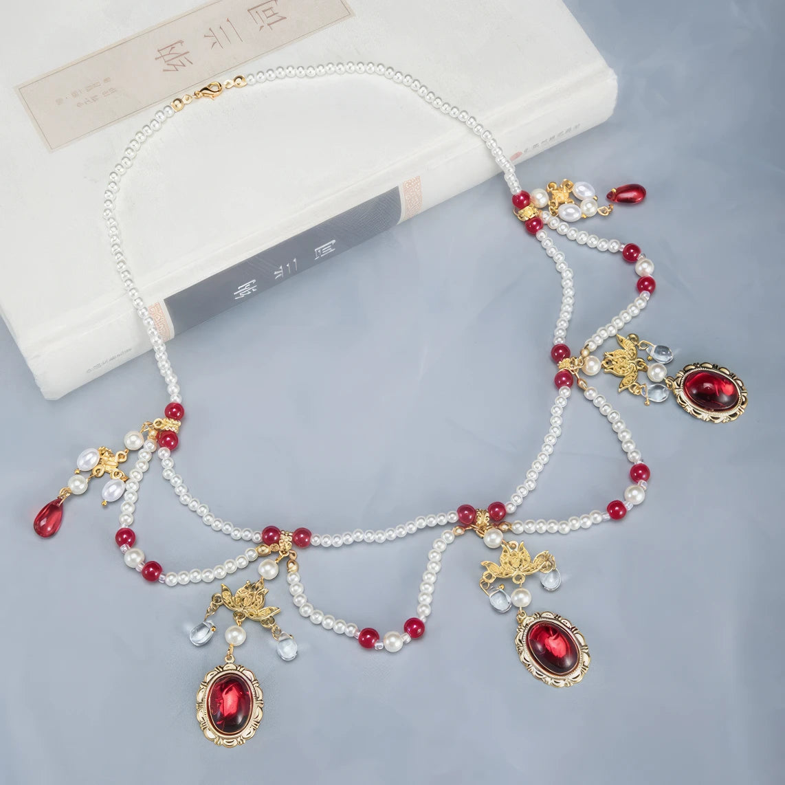 Classical pearl necklace with red beads, gold details and large red gemstones, displayed on light blue surface, book in background