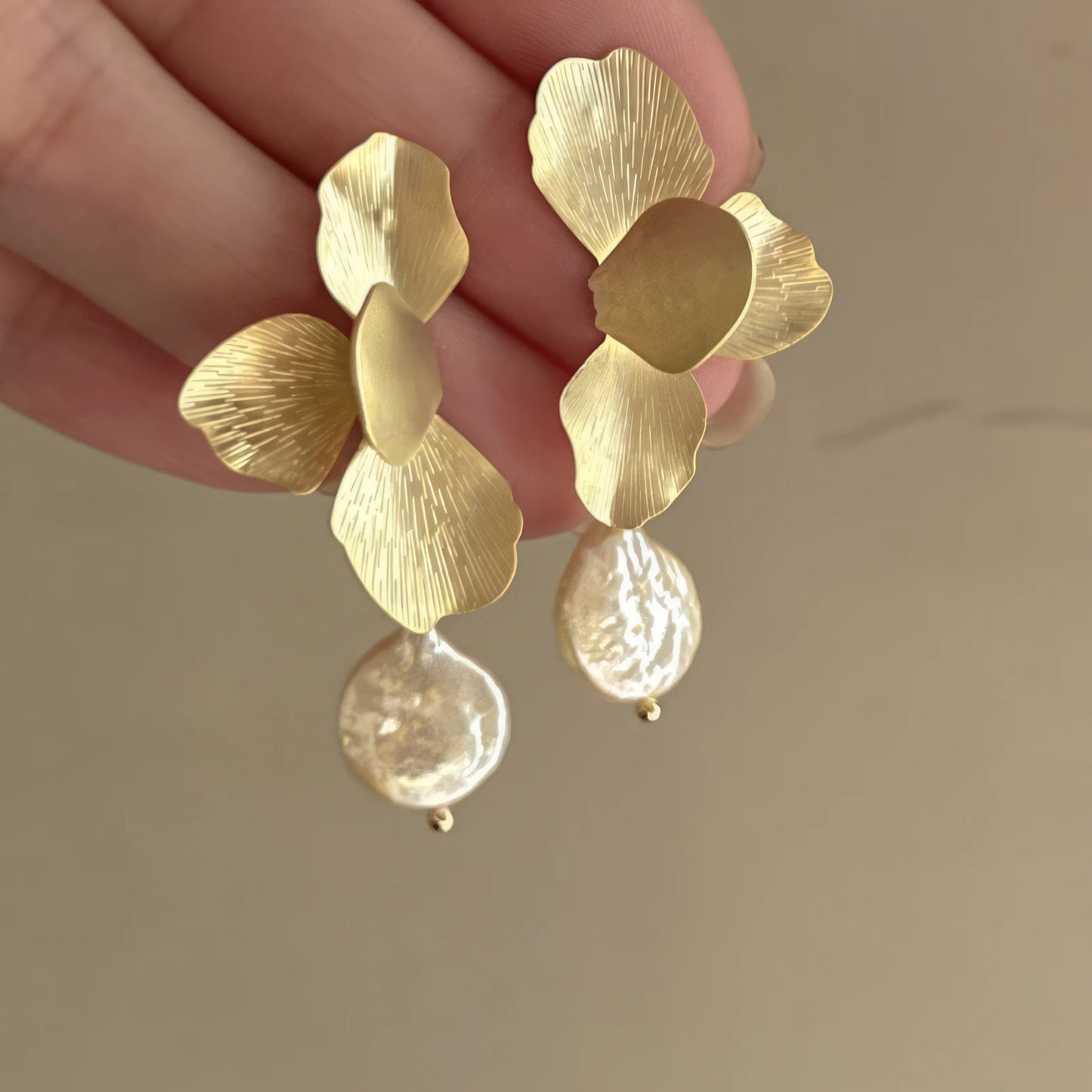 Hand holding gold flower baroque pearl earrings, showcasing design, light background