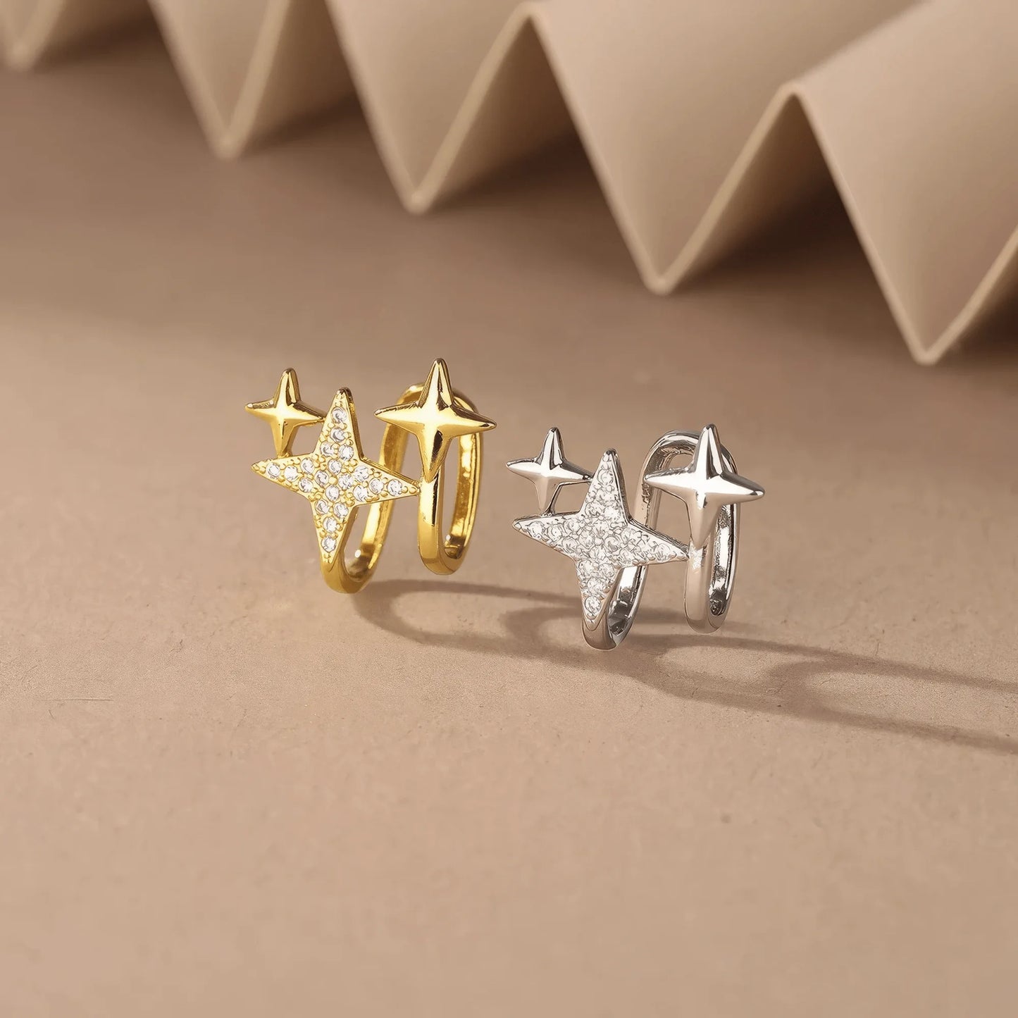 Mixed gold and silver four - point star ear clips displayed on a beige surface, emphasizing variety
