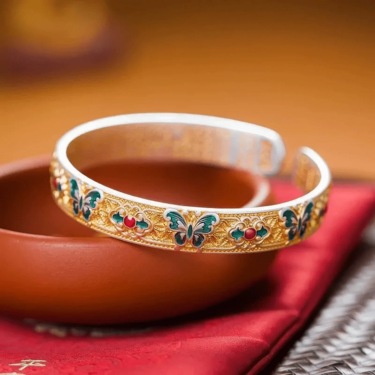 Gold - outlined cloisonné butterfly bangle displayed on red fabric with (fortune) character, emphasizing traditional theme