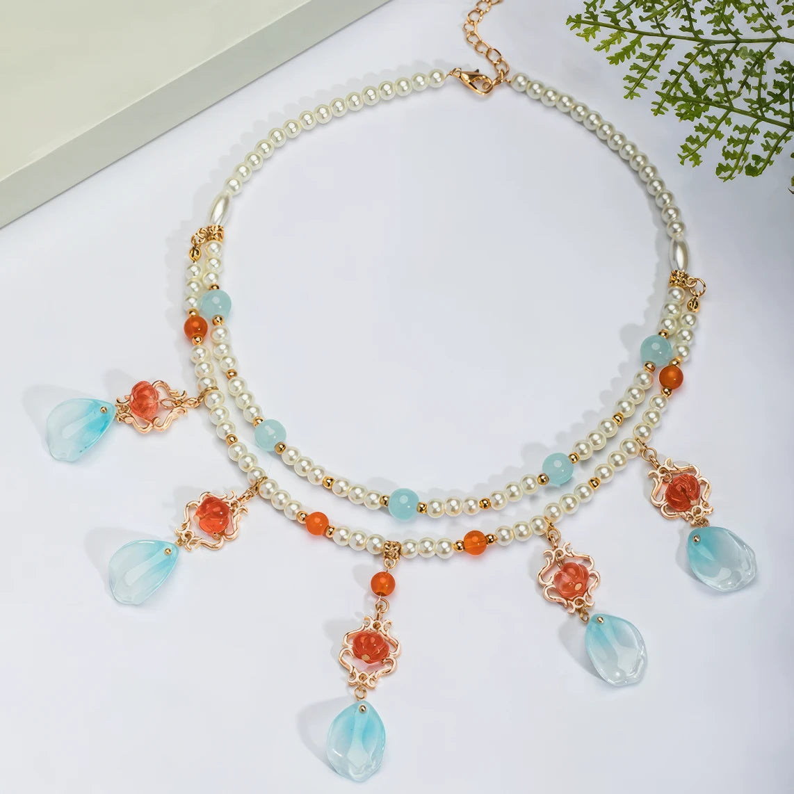 Red cotton necklace with pearls and blue beads, displayed on light surface, greenery in background