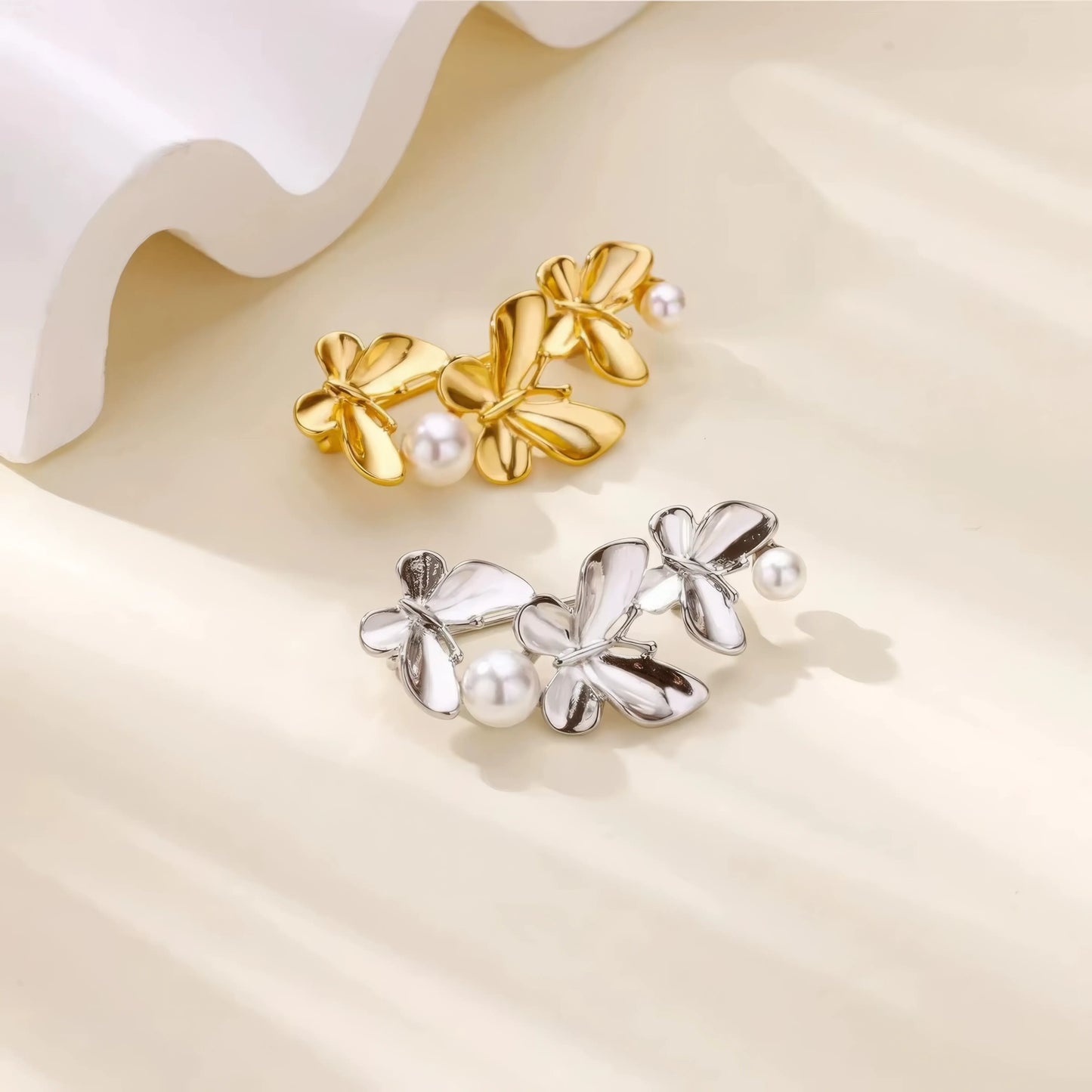 Gold and silver three - butterfly brooches with pearls, displayed on white curved surface, showcasing variety