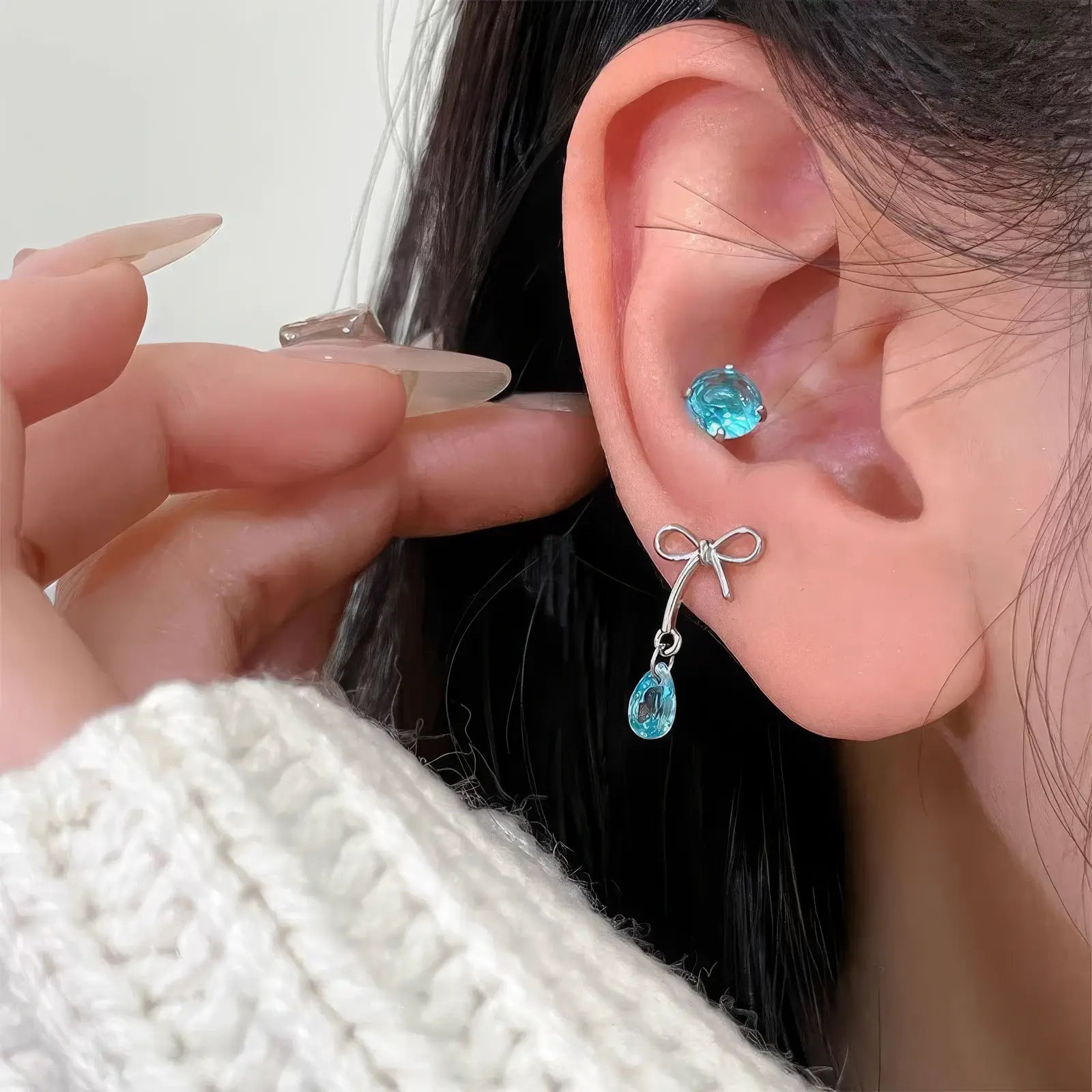 Bow - shaped blue diamond earrings worn on ear, with a hand gently touching the earring.
