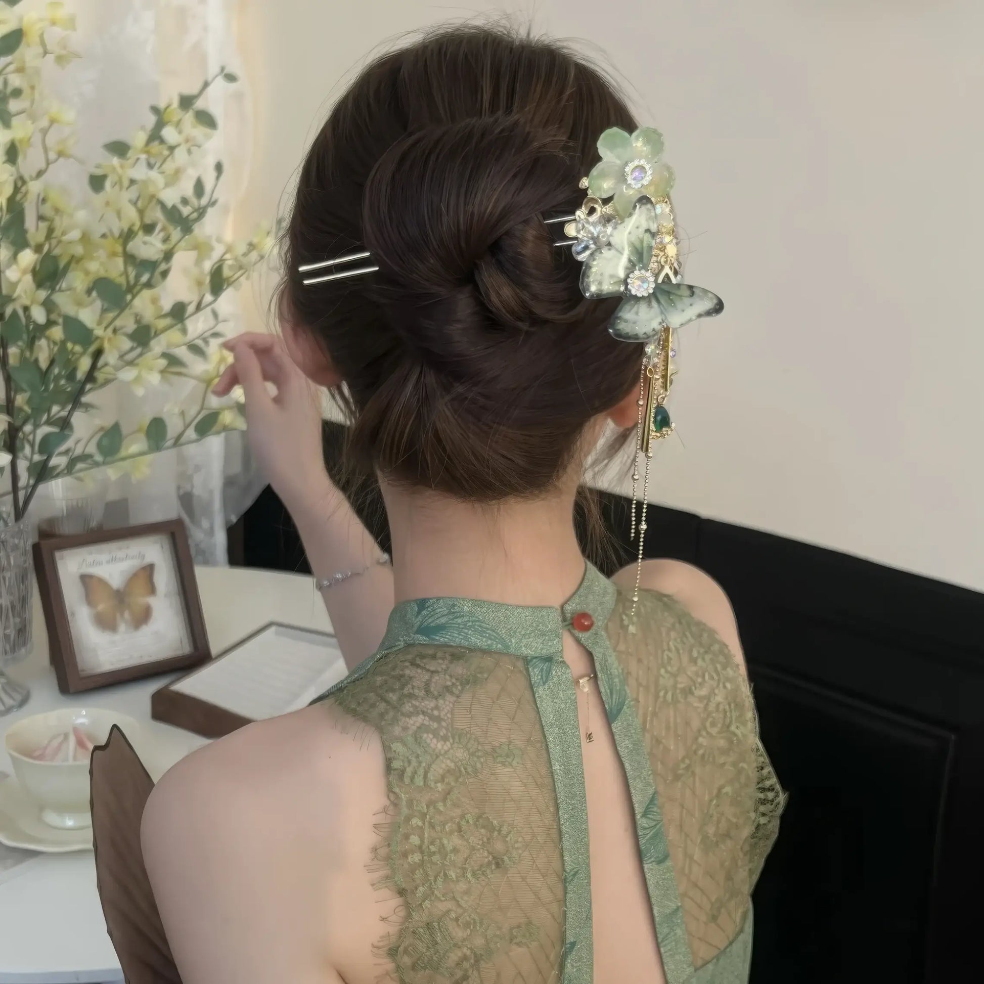 White flower butterfly hairpin secured in neat updo, wearer adjusting hair beside vintage framed decor and flowers.