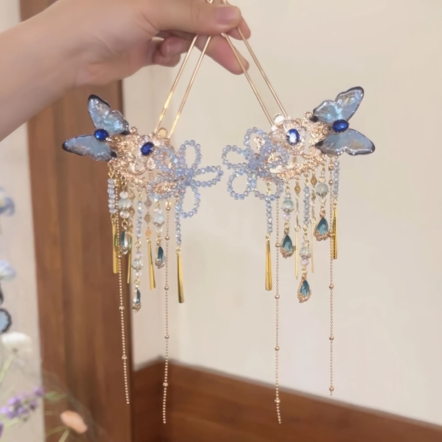 Hand gripping two blue butterfly hairpins with crystal floral design and mixed metal and stone drop tassels.