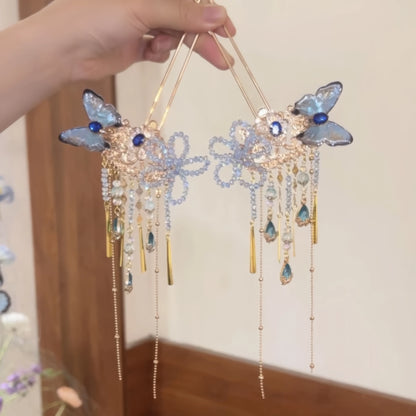 Hand gripping two blue butterfly hairpins with crystal floral design and mixed metal and stone drop tassels.