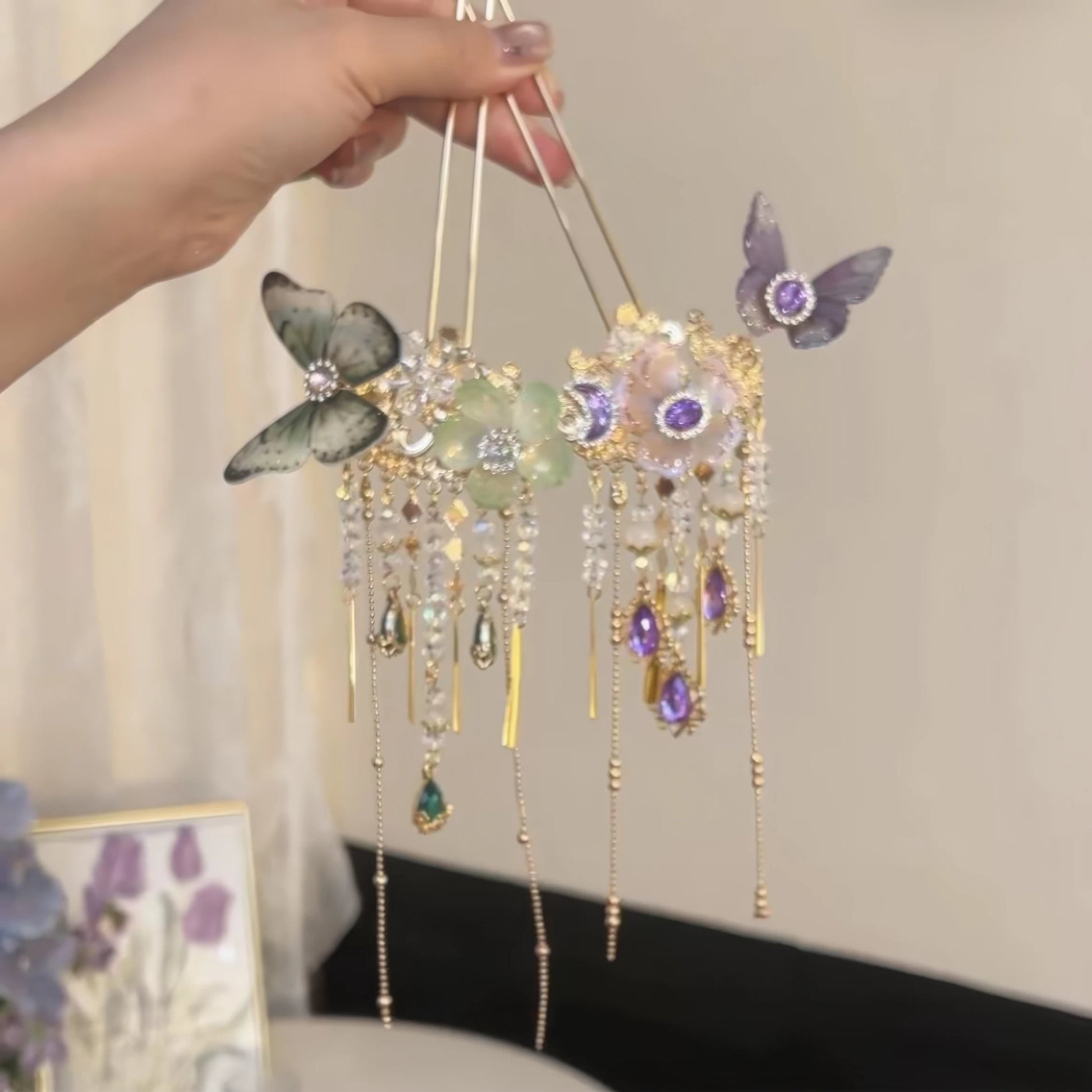Hand holding a pair of hairpins with purple and green butterfly details, dense crystal flower centerpiece and lengthy drop tassels.