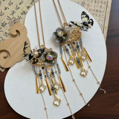 Two butterfly floral hairpins laid on antique rice paper, featuring black butterfly accents, golden pendants and dangling bead tassels.
