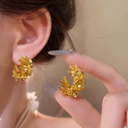 Gold floral curved earrings worn, with hand holding the other earring, showcasing the intricate, flower - clustered design