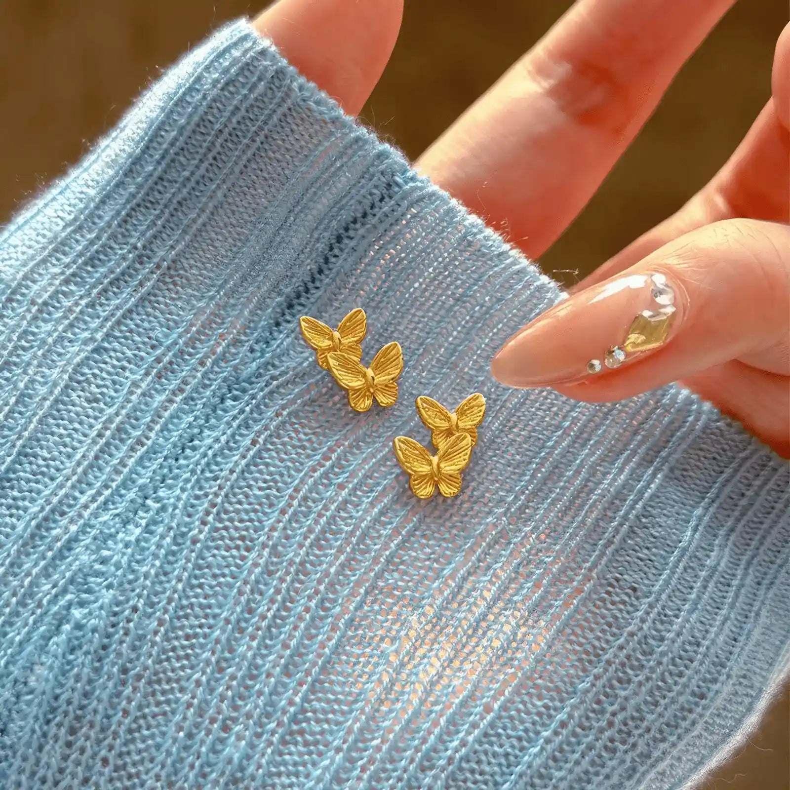 Gold butterfly stud earrings displayed on blue fabric, with hand nearby
