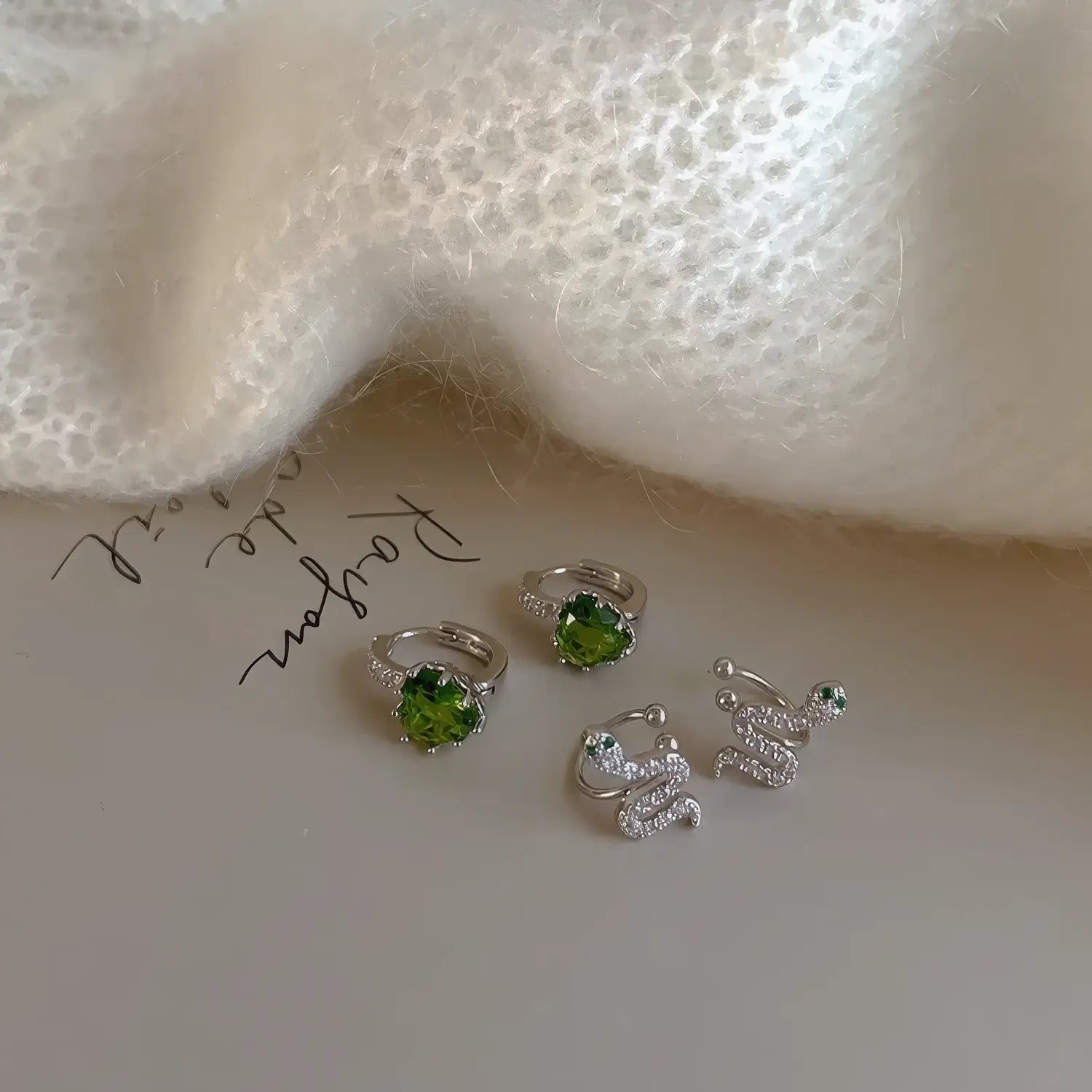 Green zircon snake - shaped stackable ear cuff along with other jewelry pieces, placed on a surface with a knitted fabric.