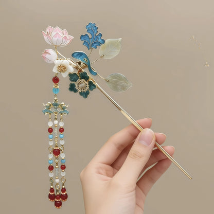 Hand holding the hairpin to show its full length, including the decorative top, pin stem, and tassel, against a neutral background.
