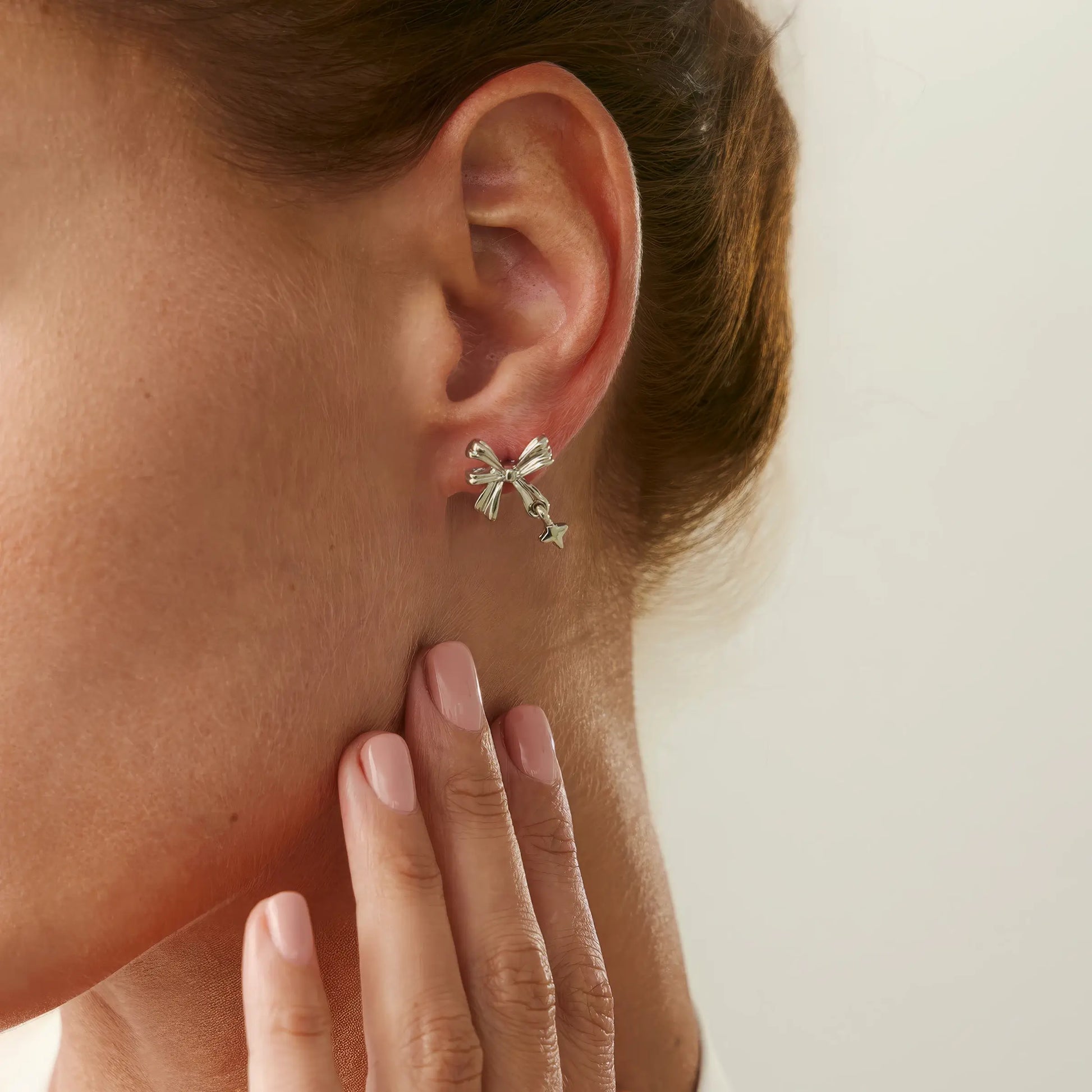 Silver bow tie stud earring worn on ear, with hand touching the ear, emphasizing the earring's design.