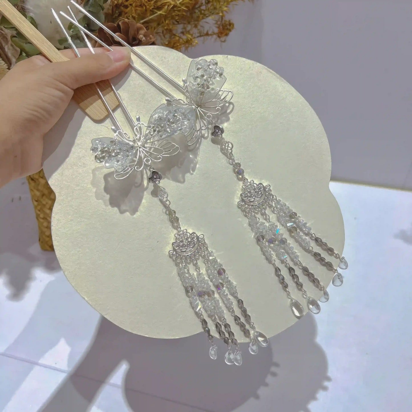 A pair of silver butterfly hairpins held on a decorative plate, showcasing the translucent butterfly design and sparkling crystal tassels.