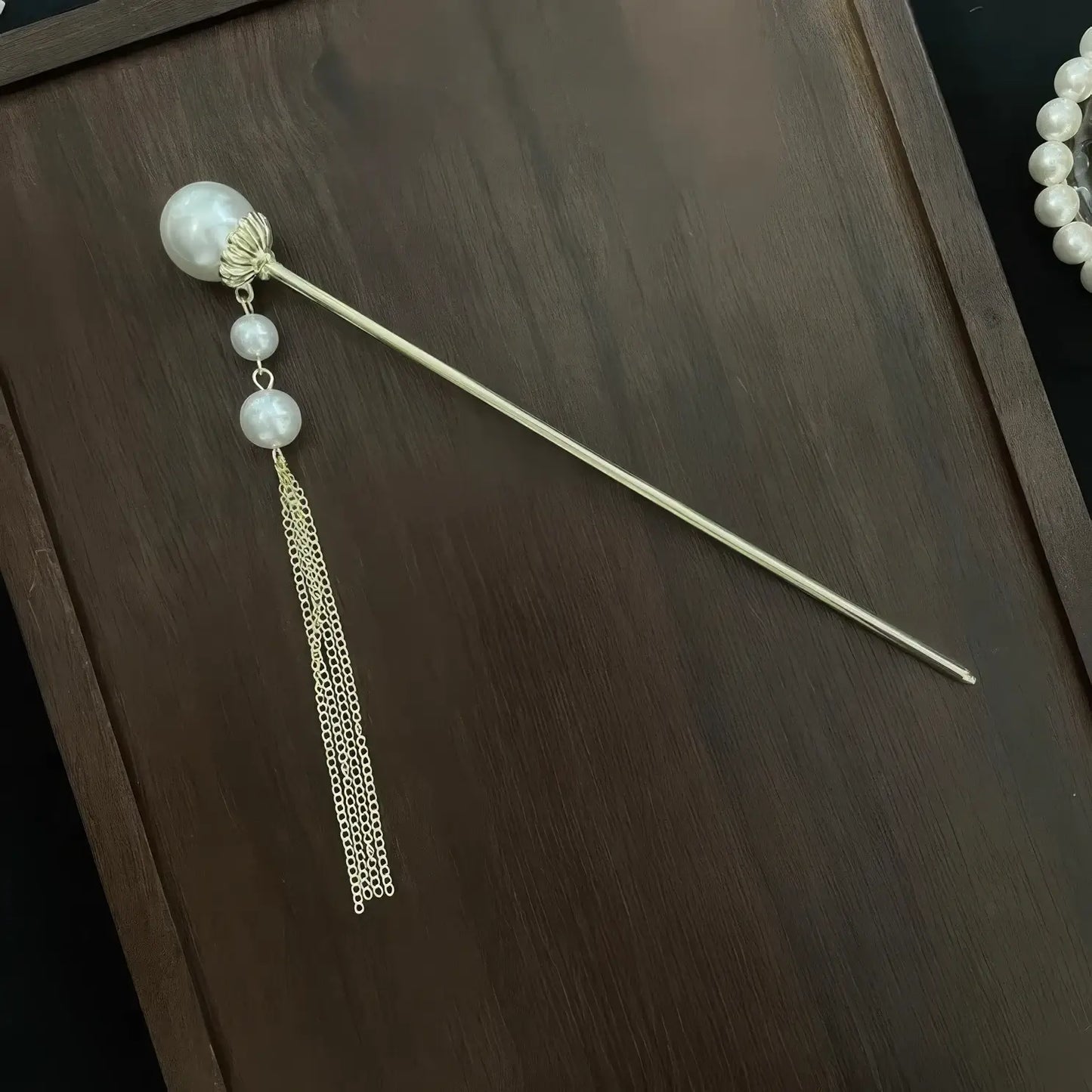 Individual golden hair stick decorated with layered pearl and thin metal tassel, placed on dark wooden board as separate matching accessory.