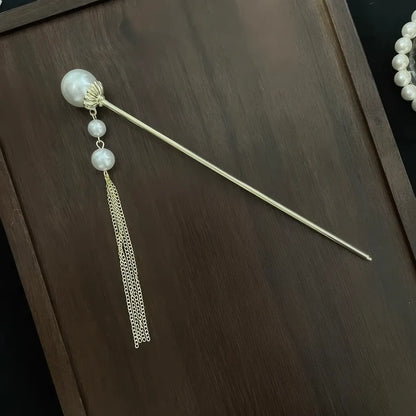 Individual golden hair stick decorated with layered pearl and thin metal tassel, placed on dark wooden board as separate matching accessory.