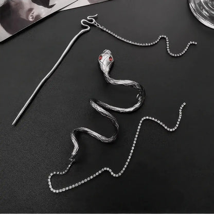 Rhinestone snake hairpin laid flat on a matte black background, showing the winding snake body and dual tassel design with full metal shine.