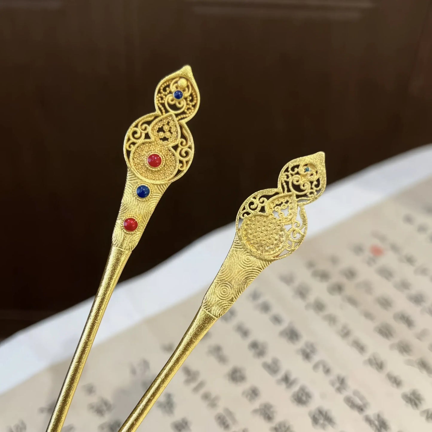 Close - up of two gold auspicious gourd hairpins with colored stones, displayed on a scroll