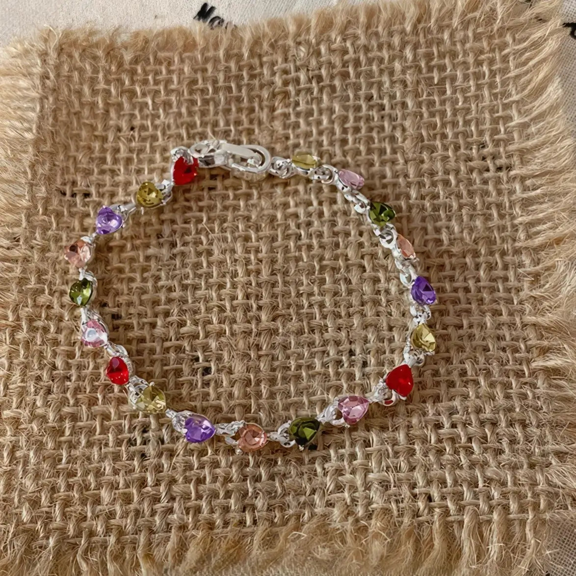 Colorful zircon heart bracelet displayed on a burlap square, beige background