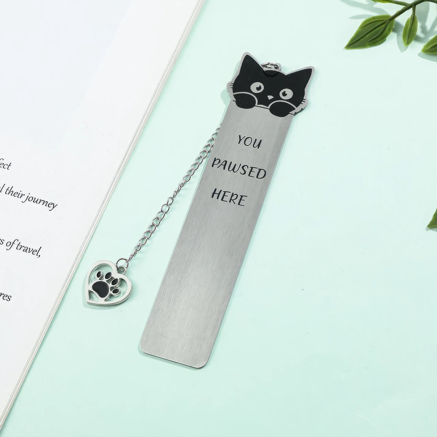 Cute cat stainless steel bookmark with chain and paw charm, displayed on green surface with leaves, white background