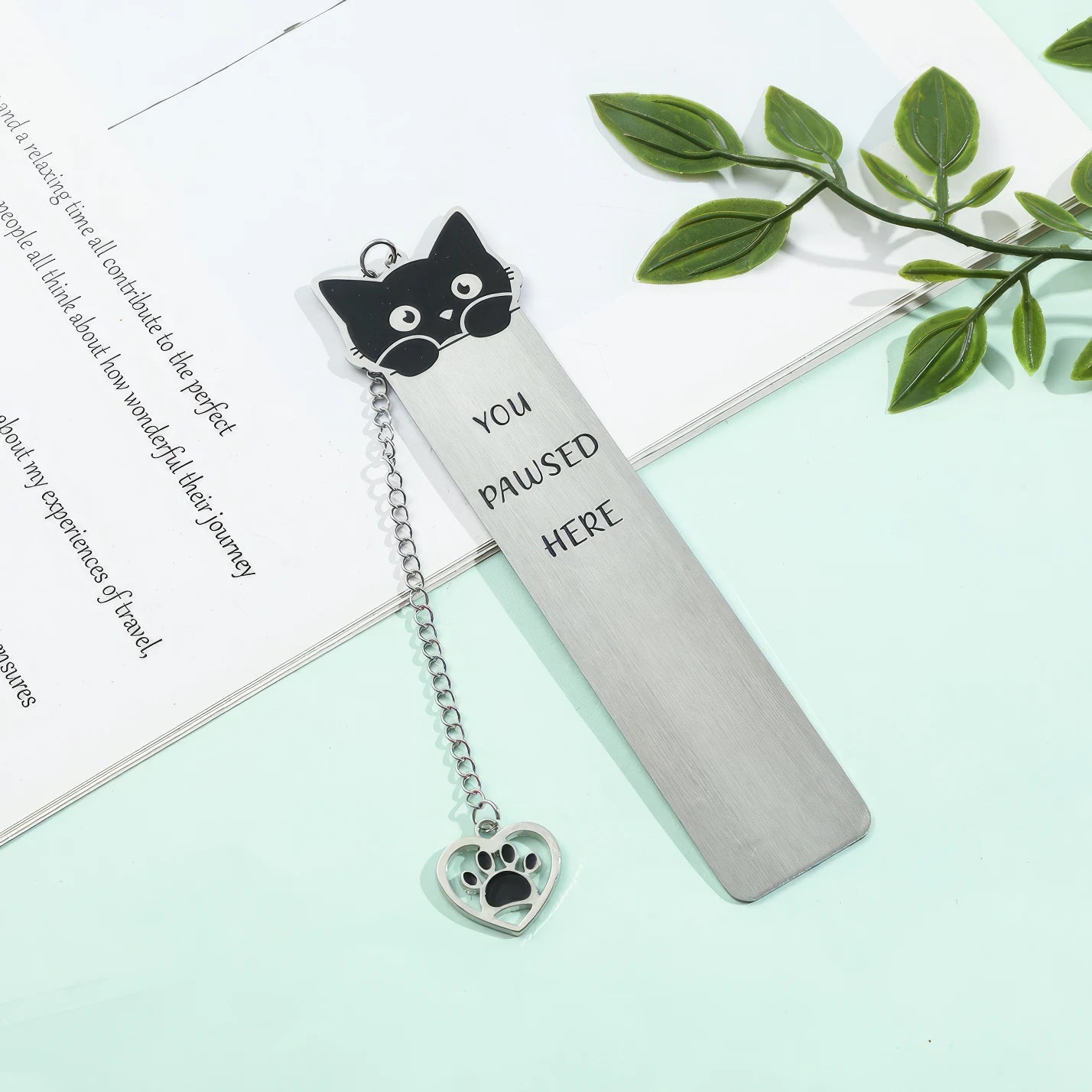 Cute cat stainless steel bookmark with chain and paw charm, displayed on green surface with leaves, white background