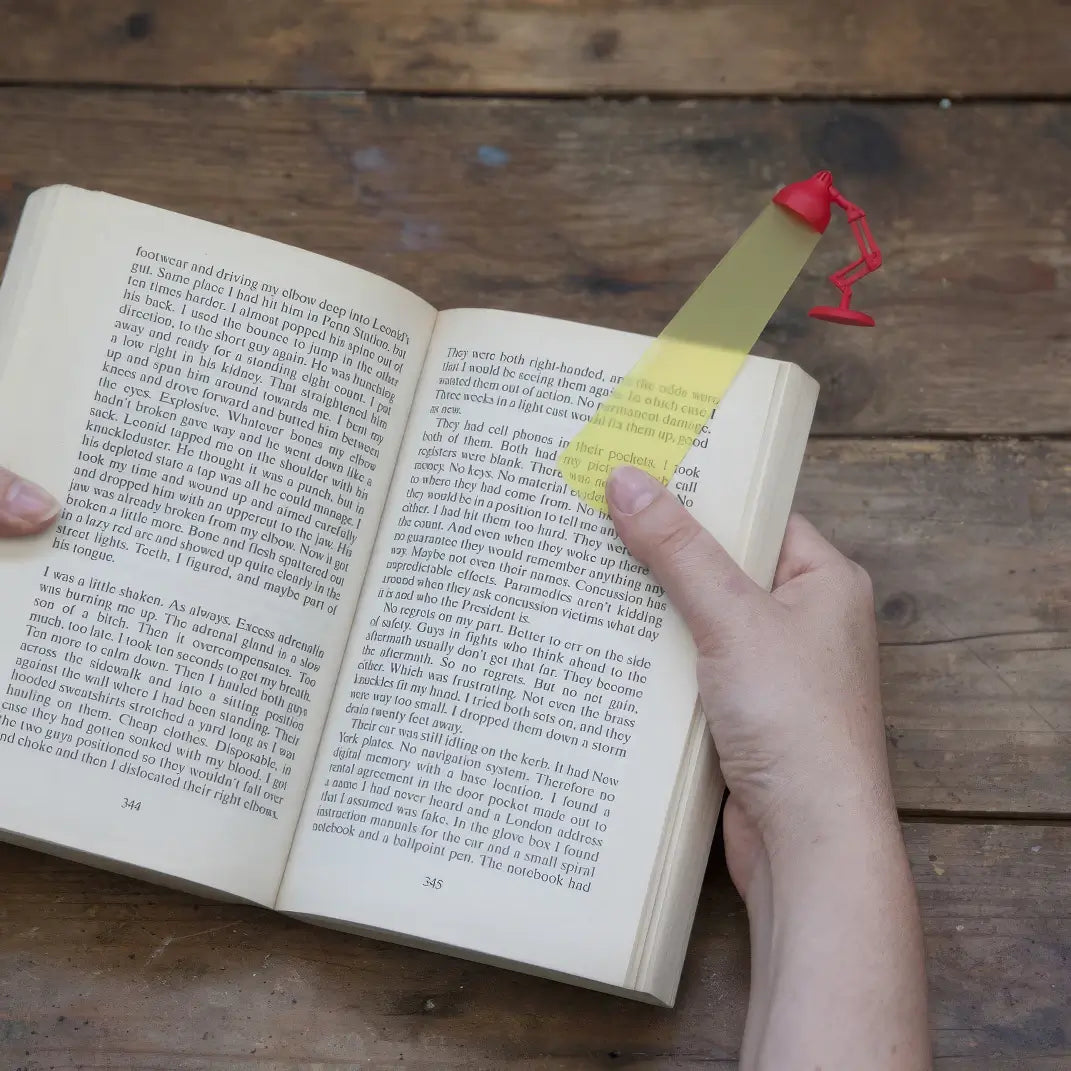 Hand using yellow - and - red desk lamp - shaped bookmark to highlight text in open book, wooden background