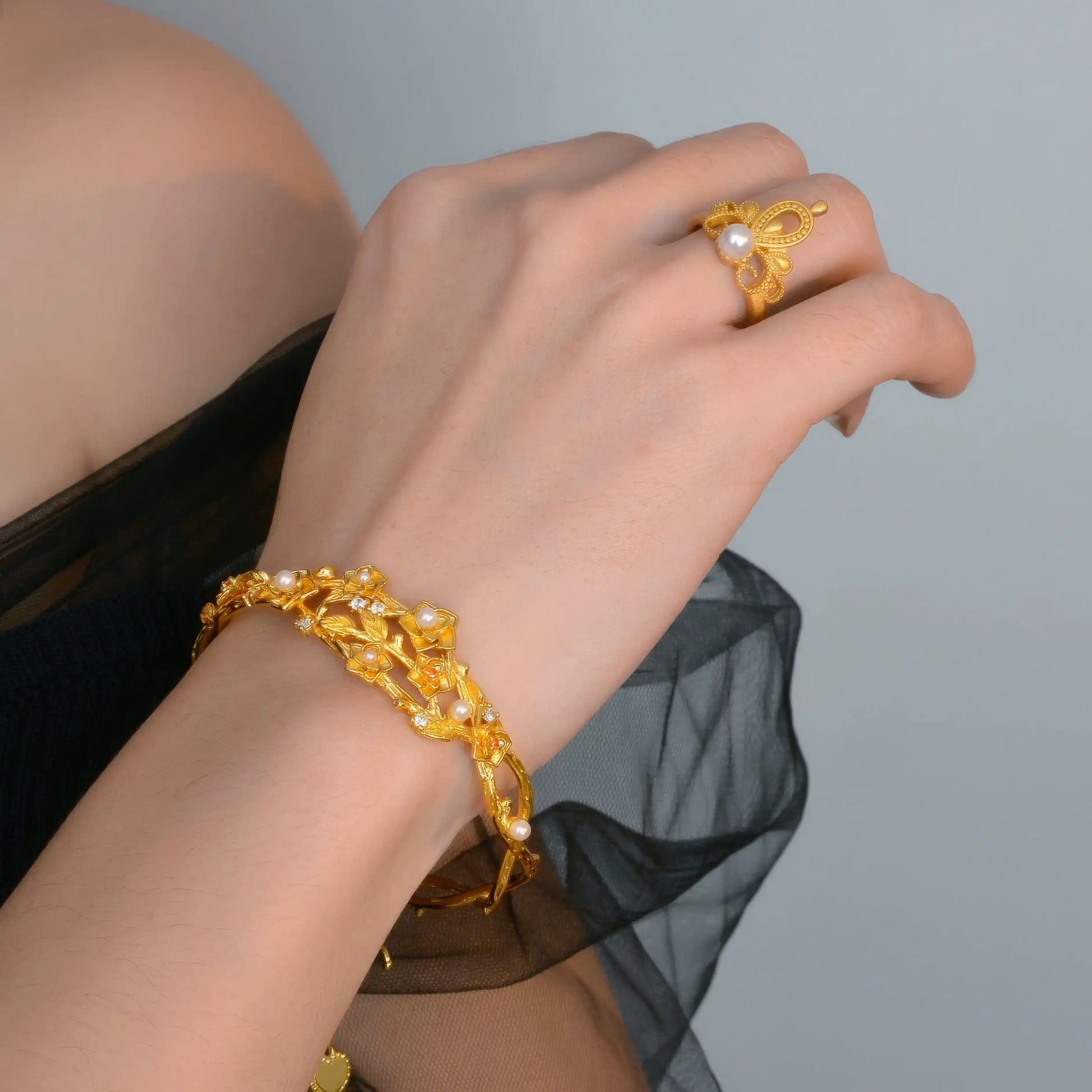 Gold flower bracelet worn on wrist, paired with ring, sheer black sleeve in background