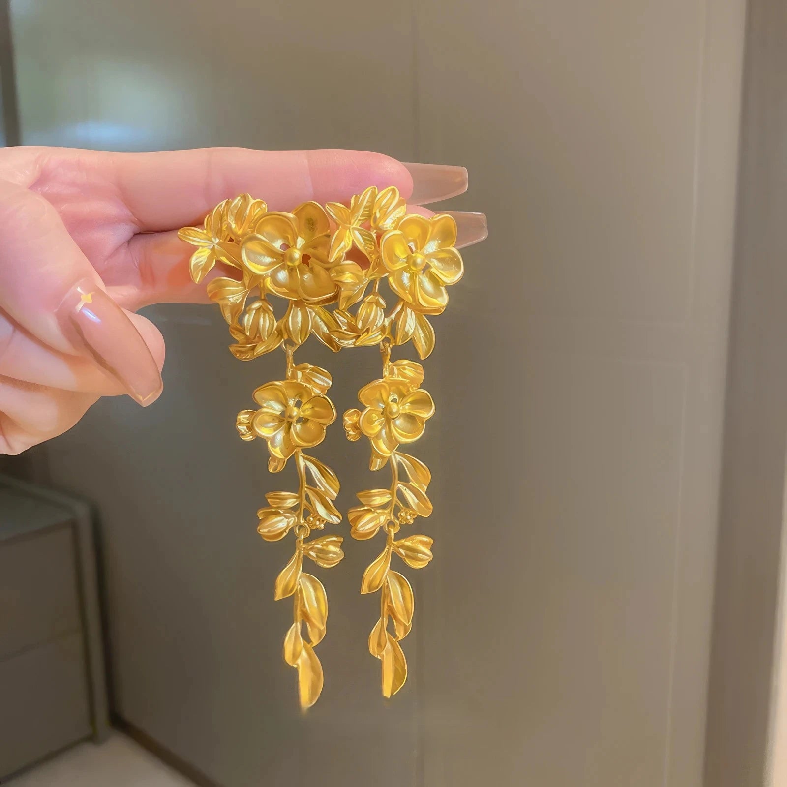Hand holding gold flower leaf tassel earrings, light background