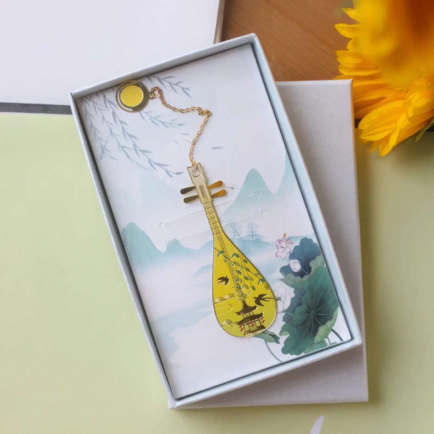 Pipa - shaped bookmark with yellow hue, presented in a white - bordered box with a scenic print, and a small chain.
