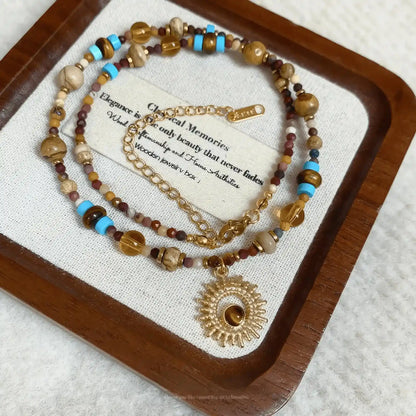 Sunflower beaded necklace, multi - color stones, gold - tone pendant, displayed on beige pad with wood and paper detail