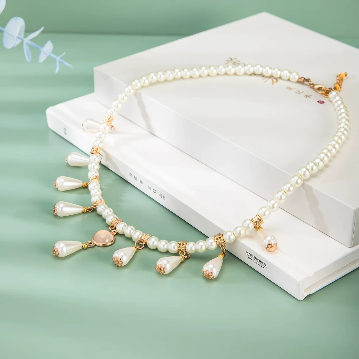 White water drop necklace with pearls and gold details, displayed on white books, green background