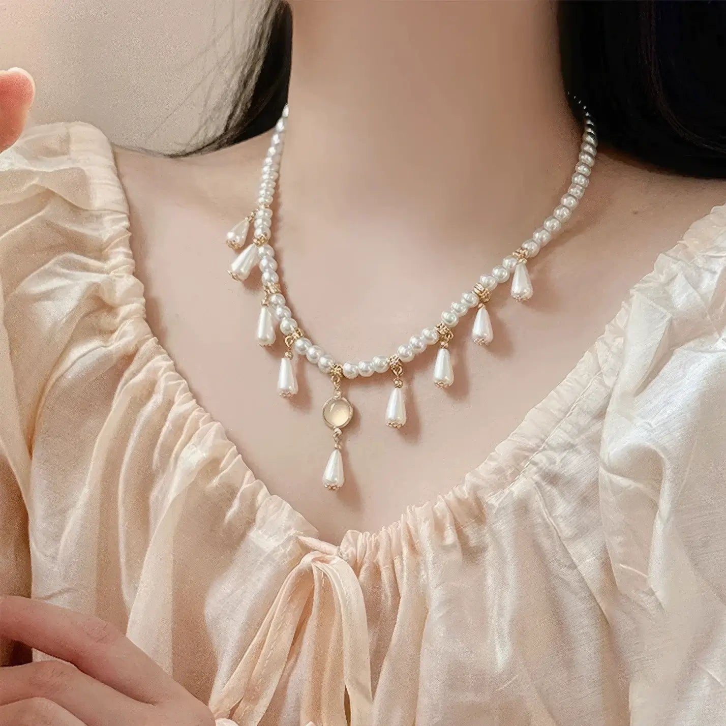 Woman wearing white water drop necklace with pearls and gold details, light - colored clothing, black hair